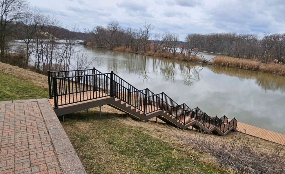 Wooden deck and staircase leading to a river, surrounded by grass and trees under a cloudy sky.