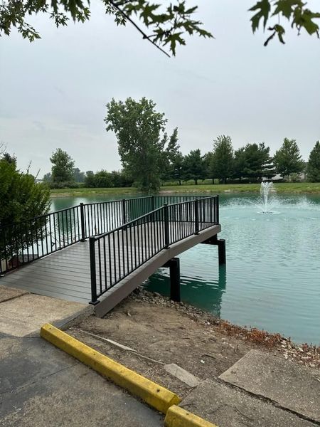 Wooden dock with black railing over turquoise water, small fountain in background, cloudy sky.
