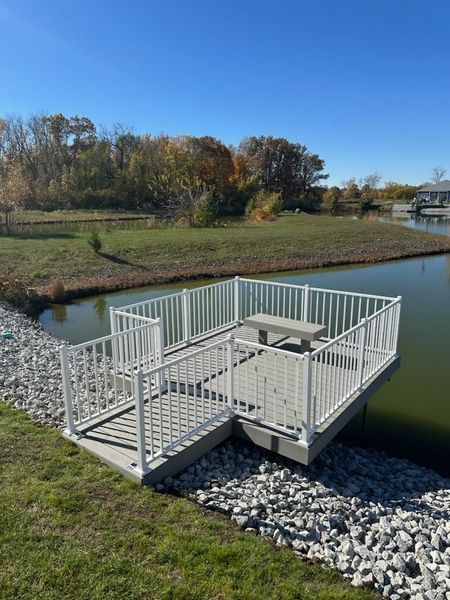 A small floating deck with white railings and a bench sits on a pond. The area is surrounded by greenery.