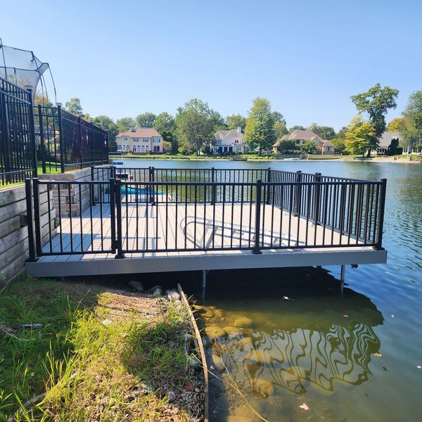 Deck over water with black railing, overlooking a lake with houses in the background. Sunny day.