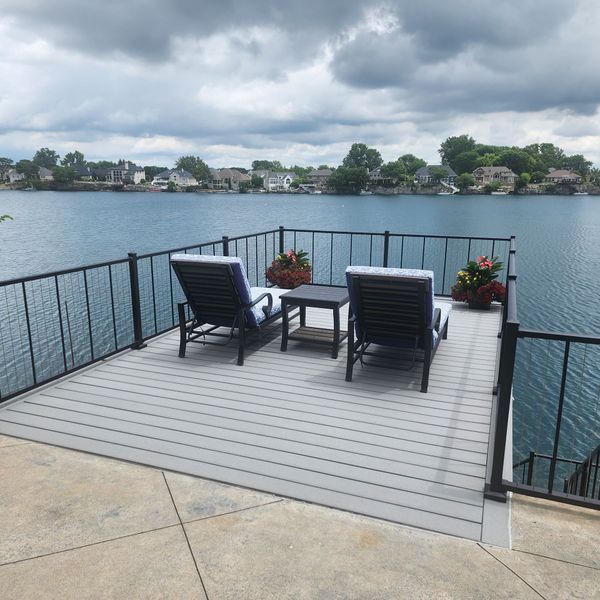 Gray deck with black railing, two lounge chairs, small table, and potted flowers overlooking a lake.