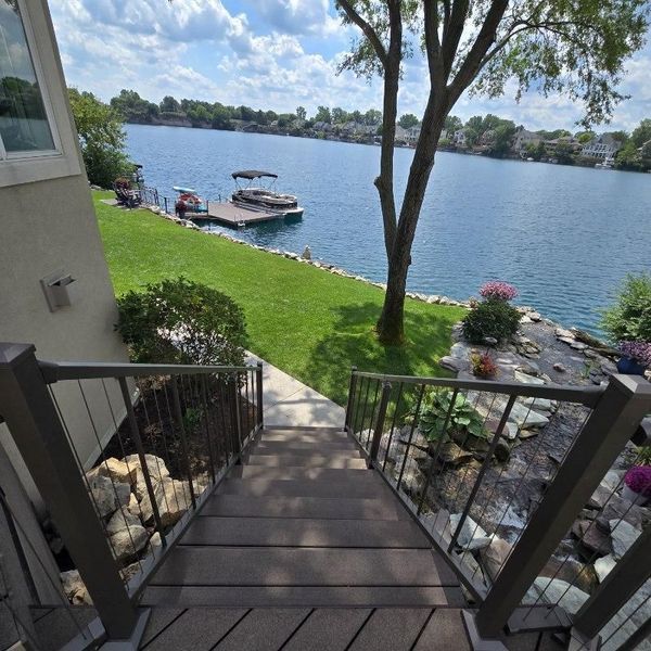 Wooden stairs lead down to a lake, dock, and green lawn. A boat is docked. Blue water and sky.