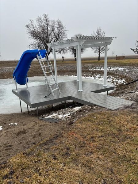 A gray dock with a blue slide, pergola, and ladder sits in a body of water in a field.