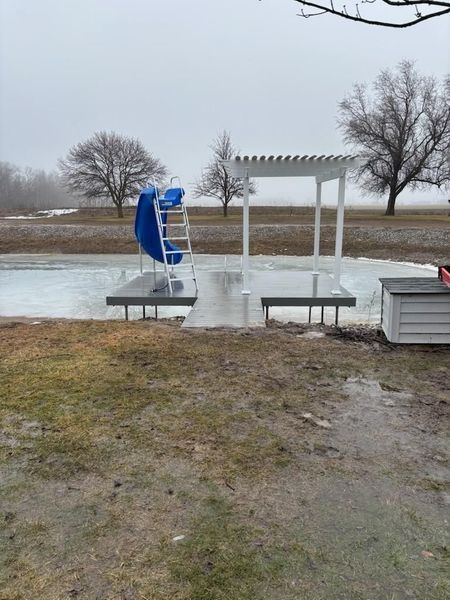 A pool area with a slide and pergola, partially covered in ice, in a field on a cloudy day.