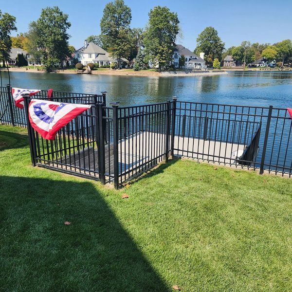 Black fence bordering a grassy yard and concrete dock at a lake. Patriotic bunting decorates the fence.