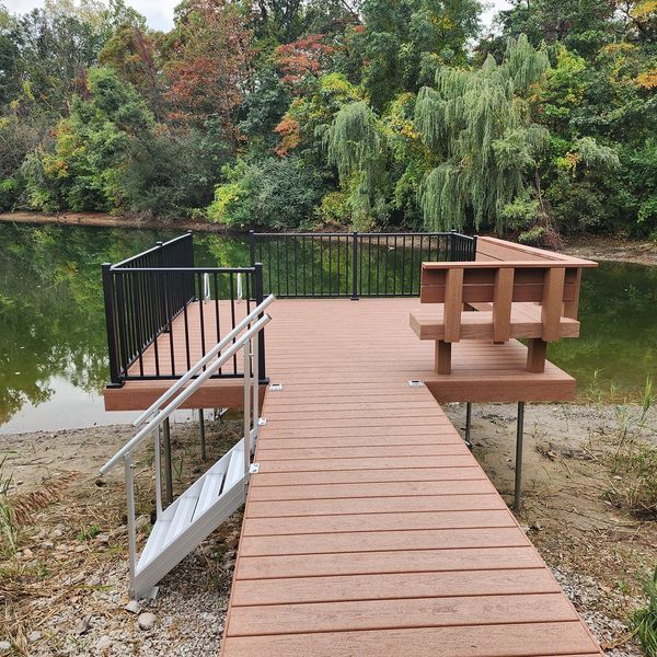 Wooden dock with black railing, bench, and ramp extending into a lake surrounded by trees.