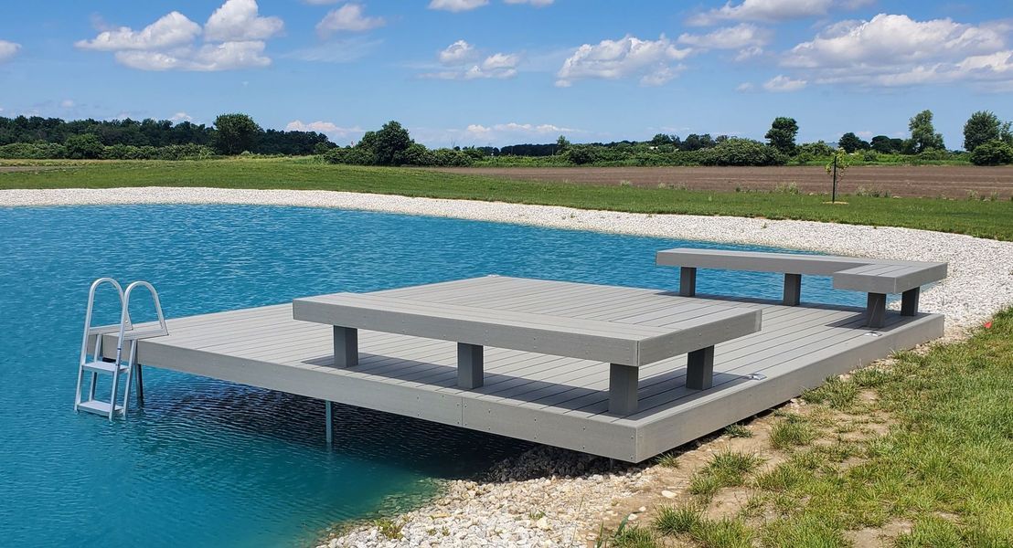 Gray dock with picnic tables and ladder in a blue pond, surrounded by grassy bank and field, blue sky.