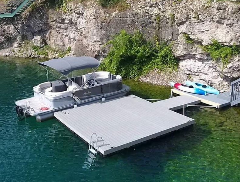 Pontoon boat and kayaks docked at a gray floating platform next to a rocky cliffside.