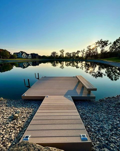 Dock extends into calm lake under blue sky; trees and houses in the background.