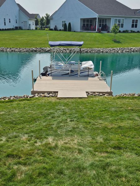 A boat docked on a wooden pier in a blue pond, surrounded by green grass and houses.