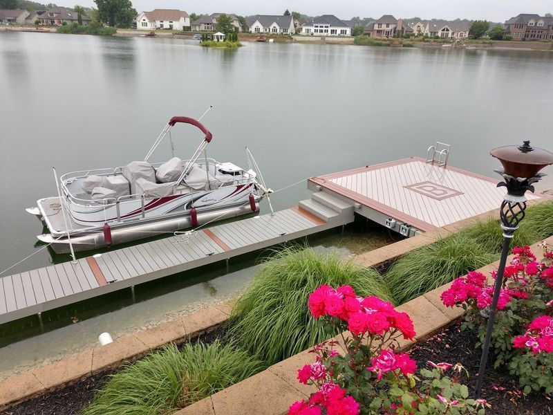Pontoon boat docked at a lakeside pier with houses in the background and pink flowers in the foreground.
