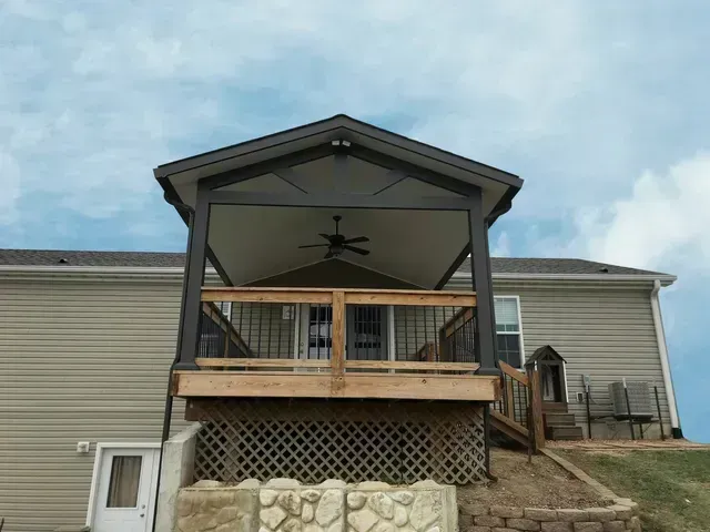 Covered back deck with a ceiling fan, attached to a light beige house under a cloudy sky.