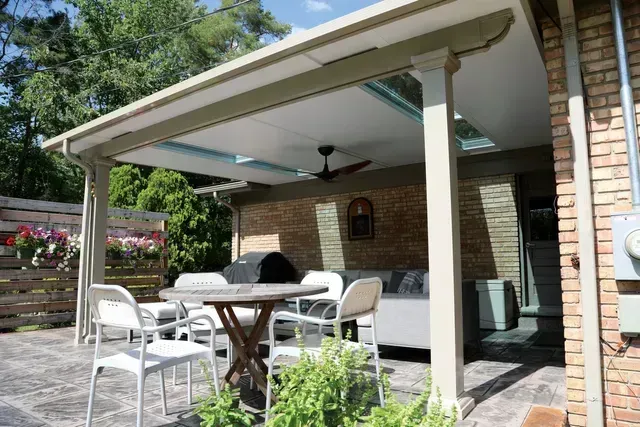 Patio with covered seating area, table, chairs, grill, and ceiling fan. Brick wall and greenery visible.