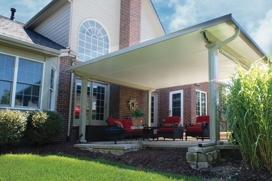 Patio with red chairs under a white roof attached to a brick house. Green lawn and tall plants.