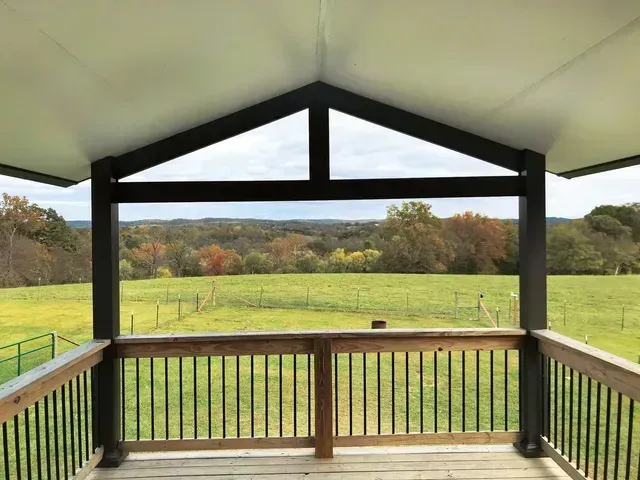 Wooden porch overlooking a green field and colorful trees. Brown railing, dark beams, and a white ceiling.