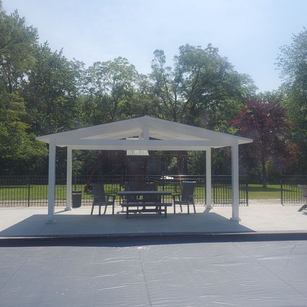 White gazebo with table and chairs on a concrete patio, trees in background, sunny day.