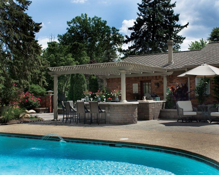 Poolside outdoor kitchen with pergola, bar seating, and surrounding trees.