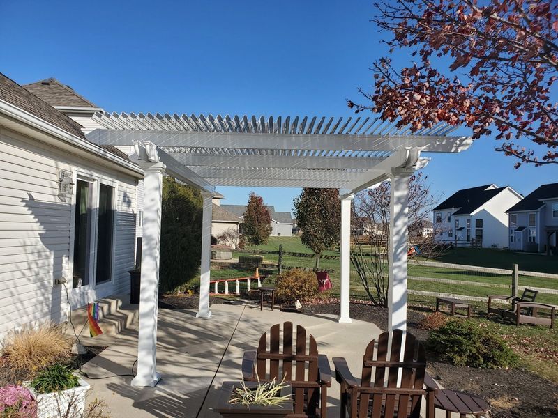 White pergola over a concrete patio with two wooden chairs. Backyard view on a sunny day.