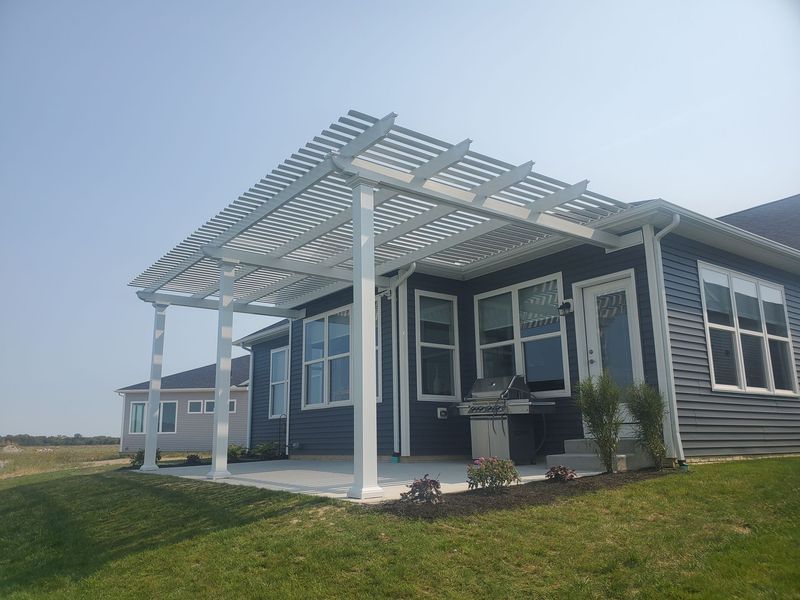 White pergola over a patio attached to a blue house with windows, grass in the foreground.