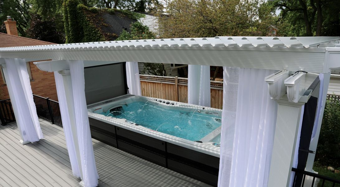 Swim spa on a deck, under a white pergola with white curtains.