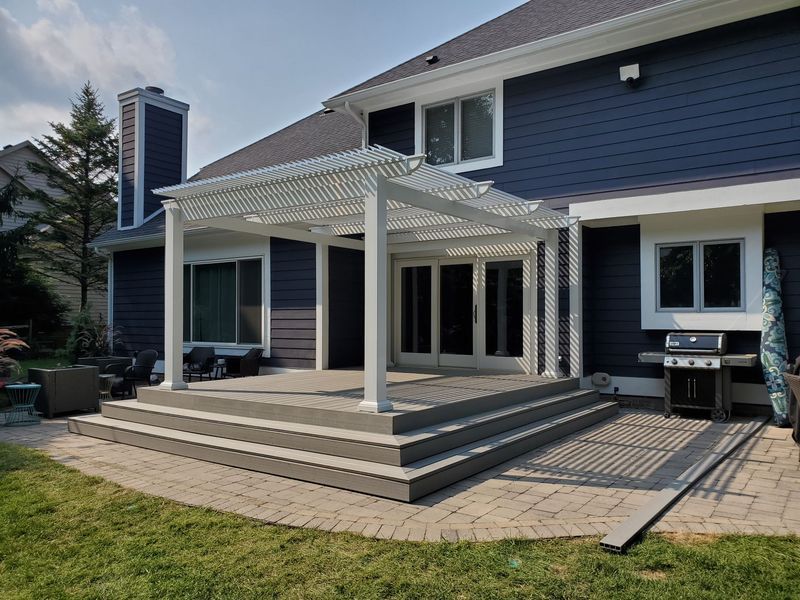 Back of a blue house with a white pergola over a deck with steps leading to a paved patio.