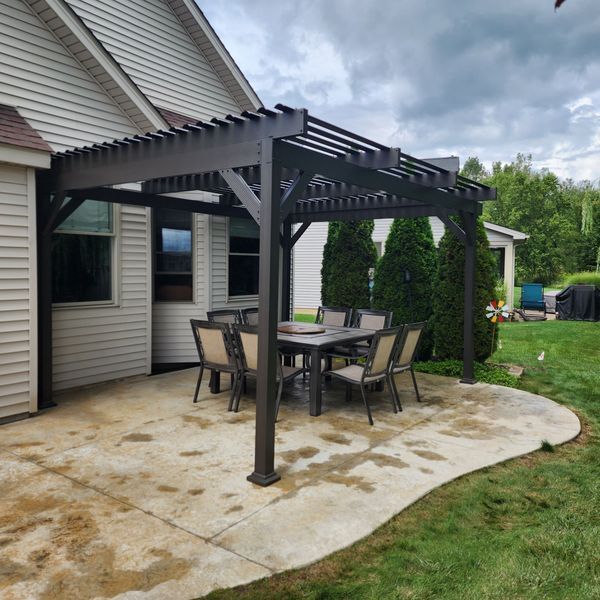 Dark pergola over a patio with a table and chairs, near a house with a green lawn.