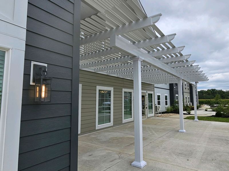 White pergola over a patio. Gray siding on a building, a lamp, and windows.