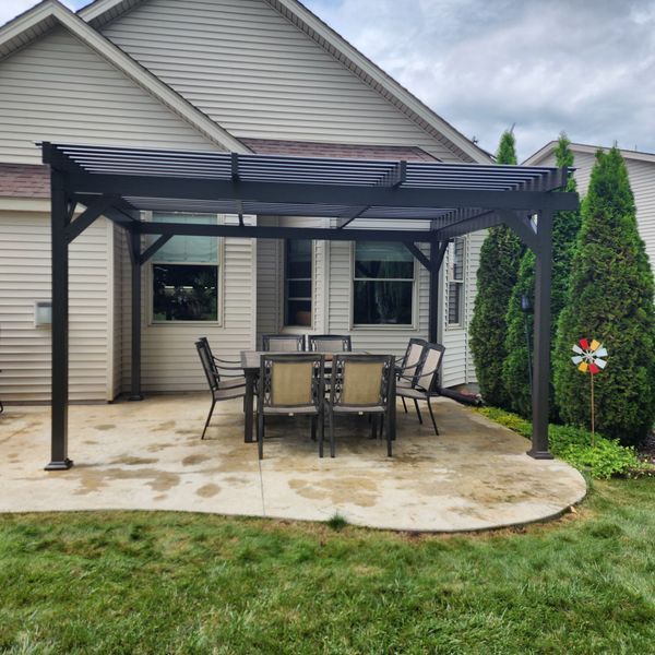 Black pergola over a patio with a table and chairs, adjacent to a house. Green grass surrounds.