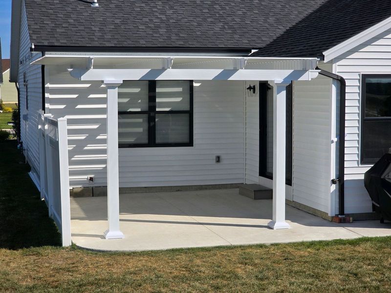 White pergola over a concrete patio outside a white house.