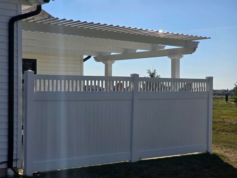 White vinyl fence next to a house with a pergola on a sunny day.
