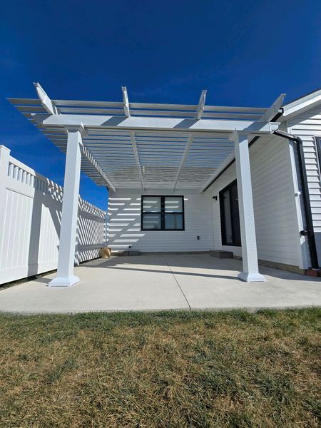 White pergola over a concrete patio outside a white house with a black window and door; sunny blue sky.