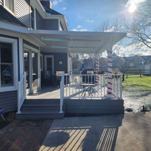 Gray and white deck with overhead cover, railing, and red/white striped pillar. Houses and yard in background.