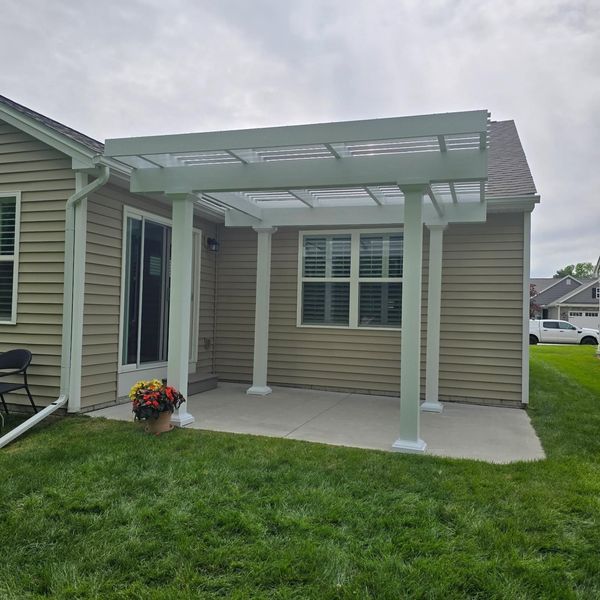 White pergola attached to a tan house with a concrete patio and green lawn.