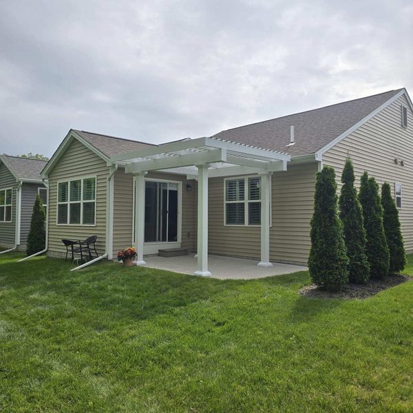 Beige house with a concrete patio and white pergola, surrounded by grass and evergreens.