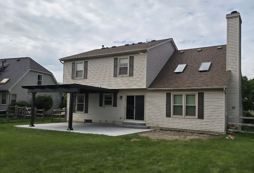 Two-story beige house with a dark pergola over a concrete patio; green lawn, cloudy sky.