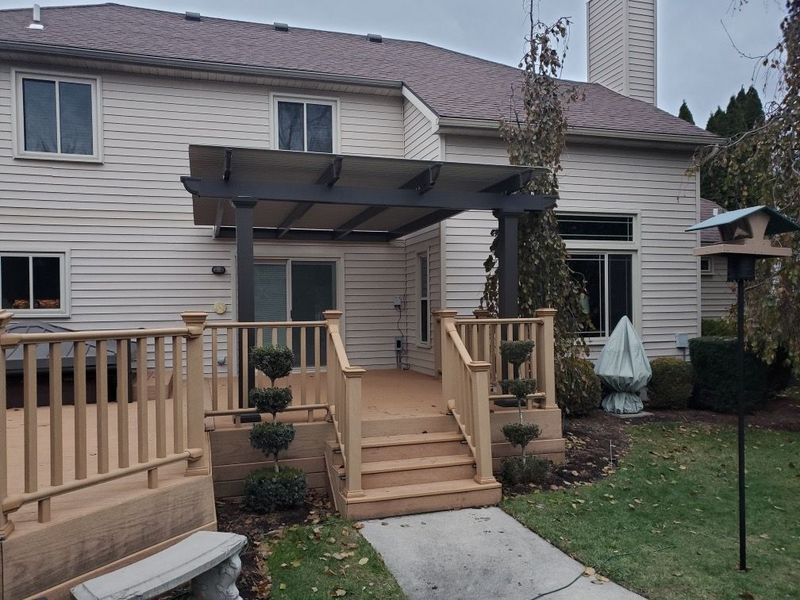 Backyard deck with pergola, steps, and a path leading to a beige house, cloudy day.