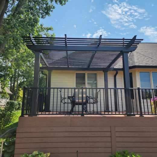 Black pergola over a deck with black railing and furniture. A house is in the background.