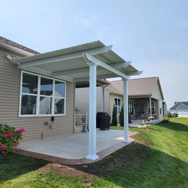 White patio cover attached to a beige house, over a concrete patio, with green grass in the foreground.
