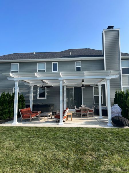 Backyard patio with white pergola, outdoor seating, and green lawn. House in background.