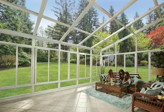 Sunroom with glass walls, overlooking a green yard. Three people sit on a wicker couch, conversing.