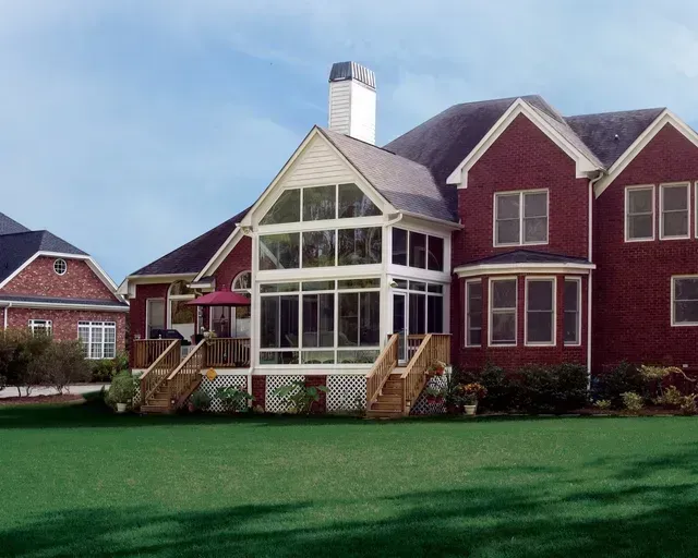 Brick house with a screened-in porch, wooden deck, and a manicured lawn under a blue sky.