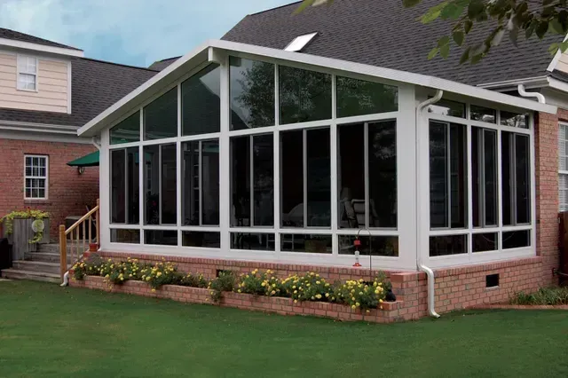 Sunroom addition with white frames, many windows, and a brick base, attached to a house with a lawn.