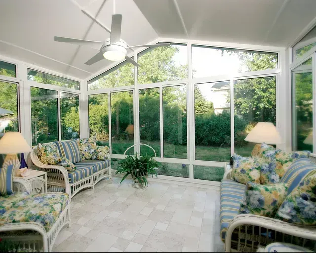 Sunroom with white wicker furniture, surrounded by large windows overlooking a green yard, ceiling fan.