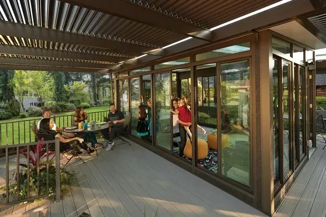 Outdoor deck with a pergola and glass-enclosed room. People are sitting at a table and inside the room.
