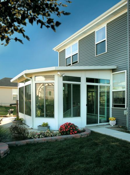 White framed sunroom addition on a gray sided house, with glass windows, and a brick lined flower bed.