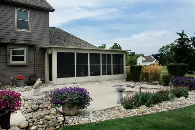 Screened porch attached to a gray house, with a concrete patio, landscaping, and a blue sky.