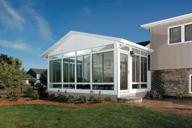 White framed glass sunroom attached to a beige house, blue sky, green lawn.