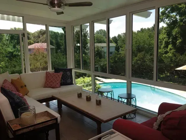 Sunroom with white sofas, coffee table, and red chair overlooking a pool and trees.