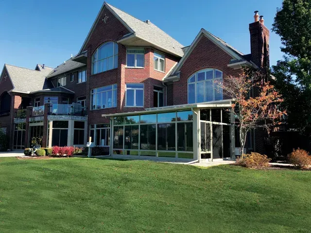 Large red brick house with a glass-walled sunroom and green lawn under a blue sky.
