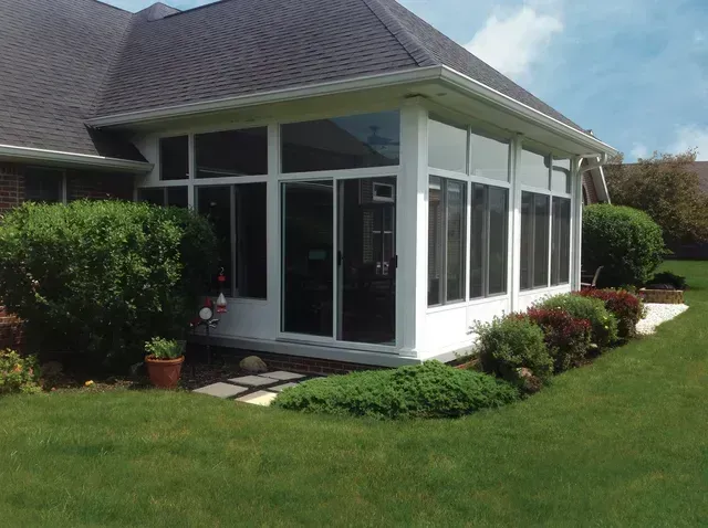 White-framed sunroom with large windows attached to a brick home. Lush green bushes surround the room in a grassy yard.